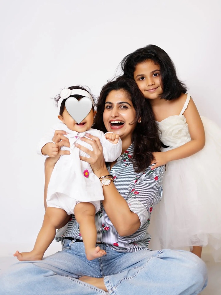 Chaithra Rai sitting on the floor holding her baby daughter, her elder daughter leaning over her shoulder