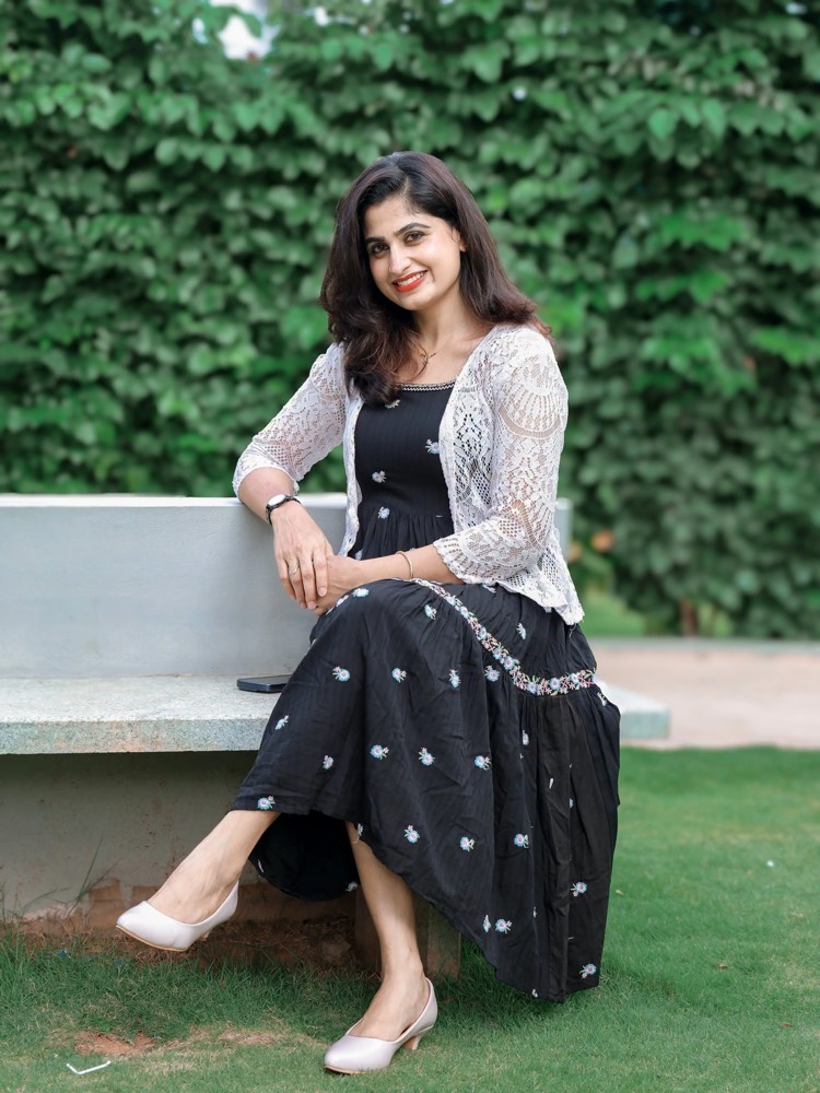 Chaithra Rai seated on a stone bench in a black embroidered dress and white lace shrug, against a wall of creeper