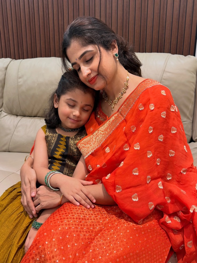 Chaithra Rai in an orange saree with her elder daughter asleep against her shoulder