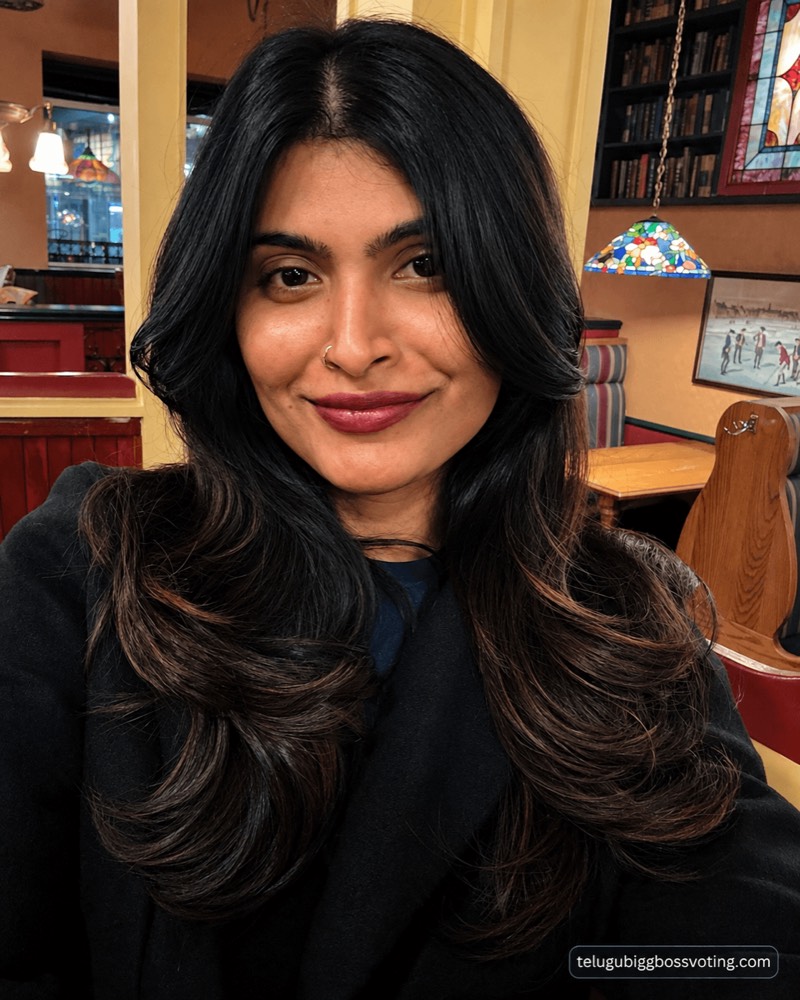 Illustrated selfie-style portrait of Gayathri Vardhan in a black coat inside a warm-lit cafe with stained-glass lamps