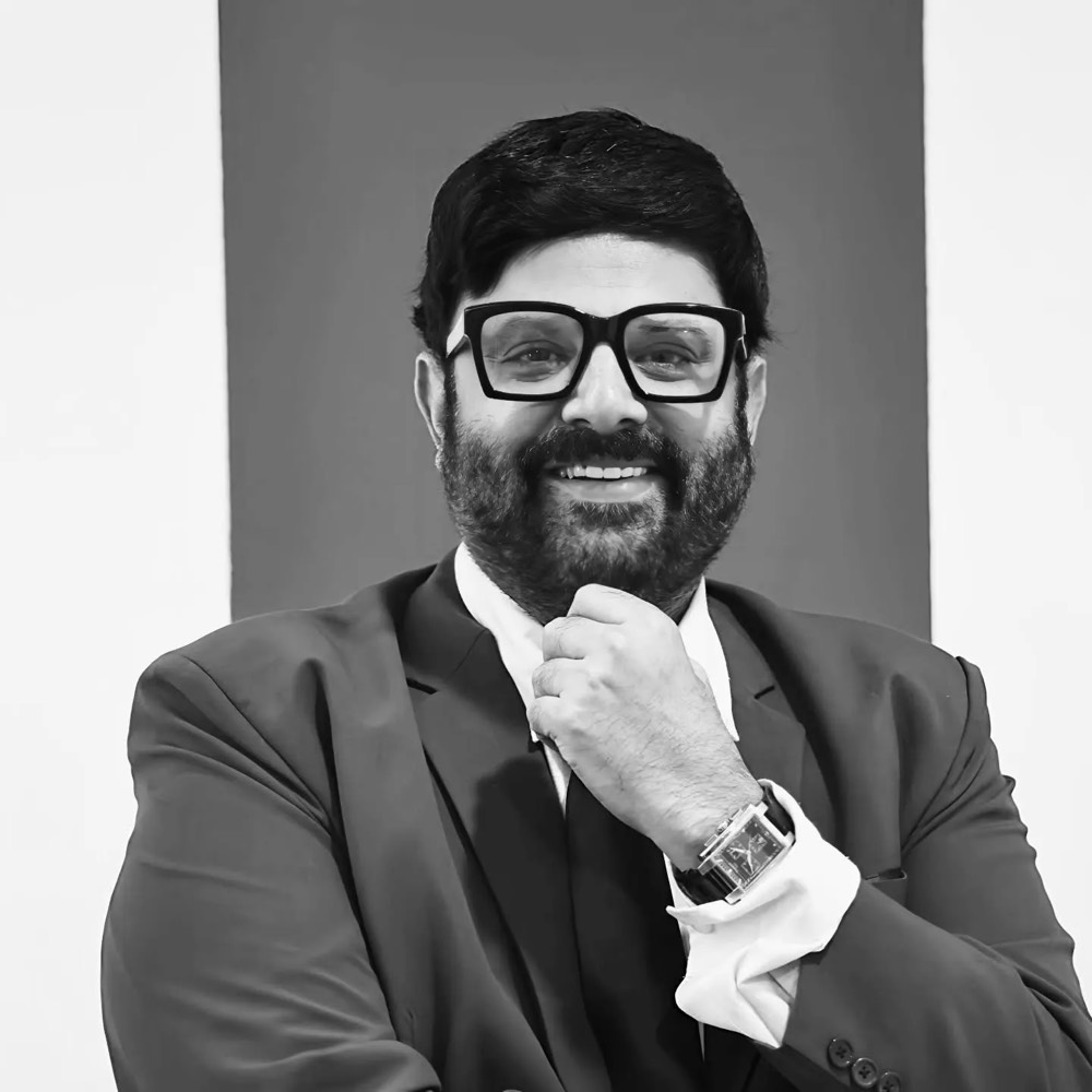 Black and white studio portrait of Krishnudu in a three-piece suit and heavy black-framed glasses, hand at his collar