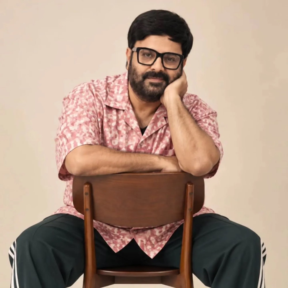 Krishnudu sitting astride a wooden chair in a pink printed shirt and dark trousers, chin resting on his hand, against a plain beige backdrop