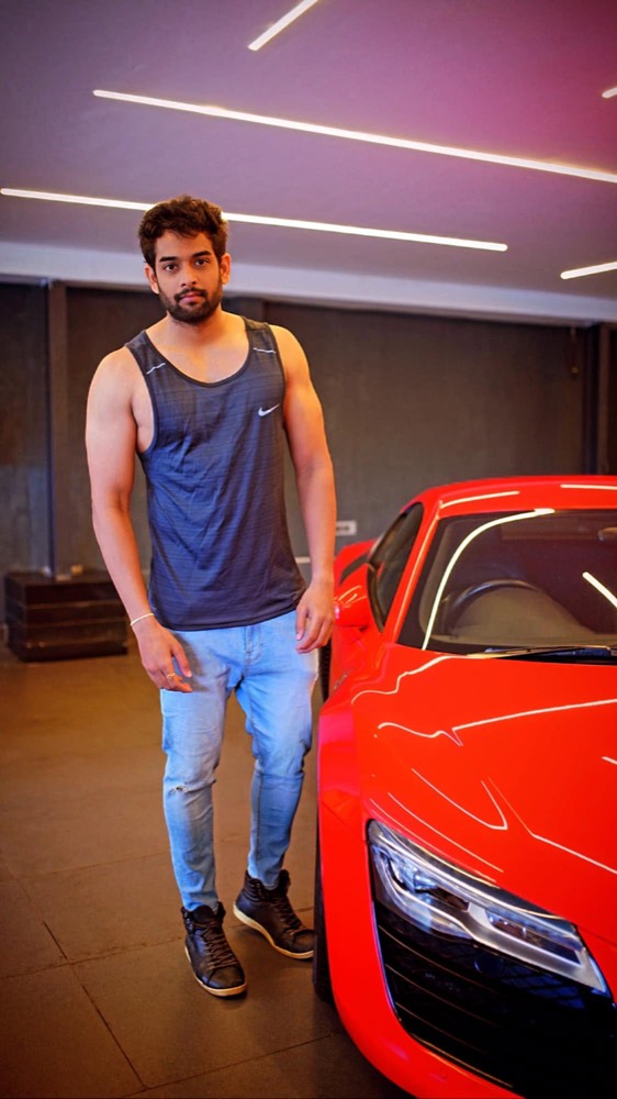 Sudheer Reddy standing beside a red sports car in a showroom, in a sports vest and jeans