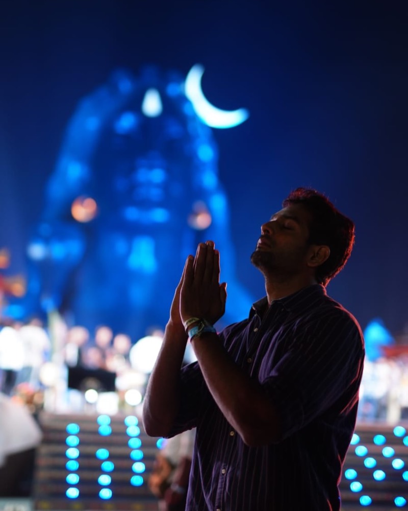 Sudheer Reddy standing with his palms joined in front of the illuminated Adiyogi statue at night