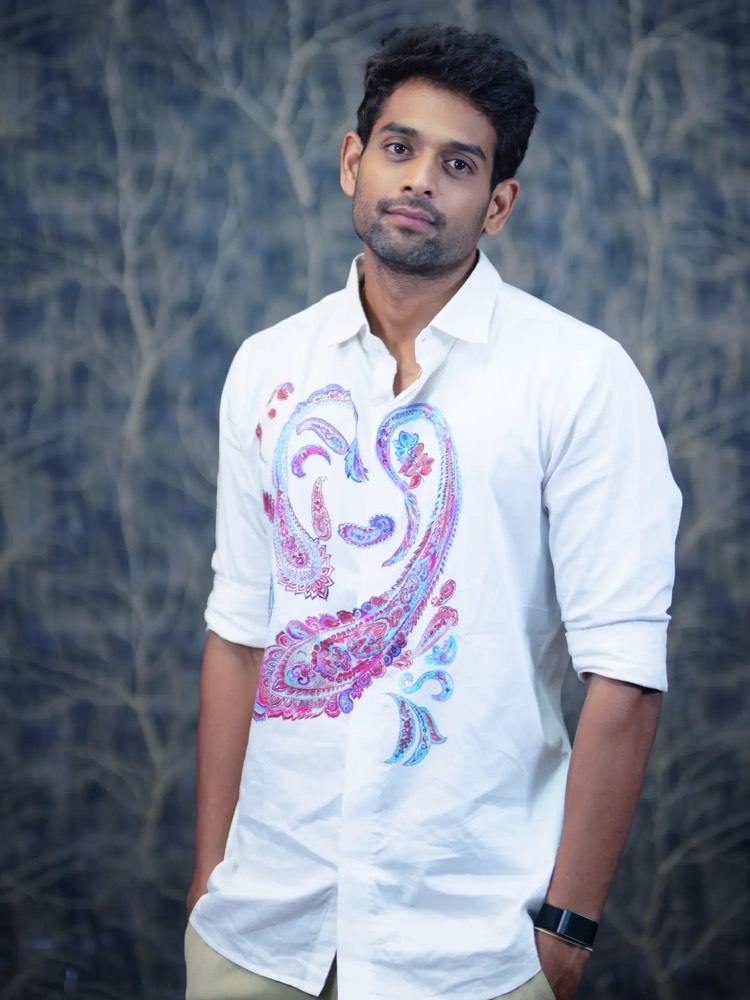 Sudheer Reddy standing in a white shirt with a large paisley print, against a dark painted backdrop