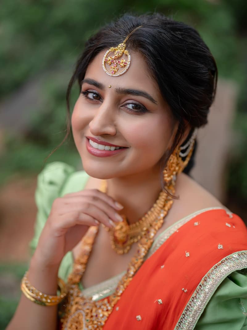 Varshini Sounderajan in an orange and green half saree with temple jewellery, smiling